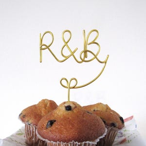 Könnte beinhalten: Ein goldener Draht-Kuchenaufsatz mit den Buchstaben "R&B" über drei Muffins. Die Muffins sind goldbraun mit dunklen Flecken, wahrscheinlich Blaubeeren, und in Papierförmchen. Der Hintergrund ist weiß.