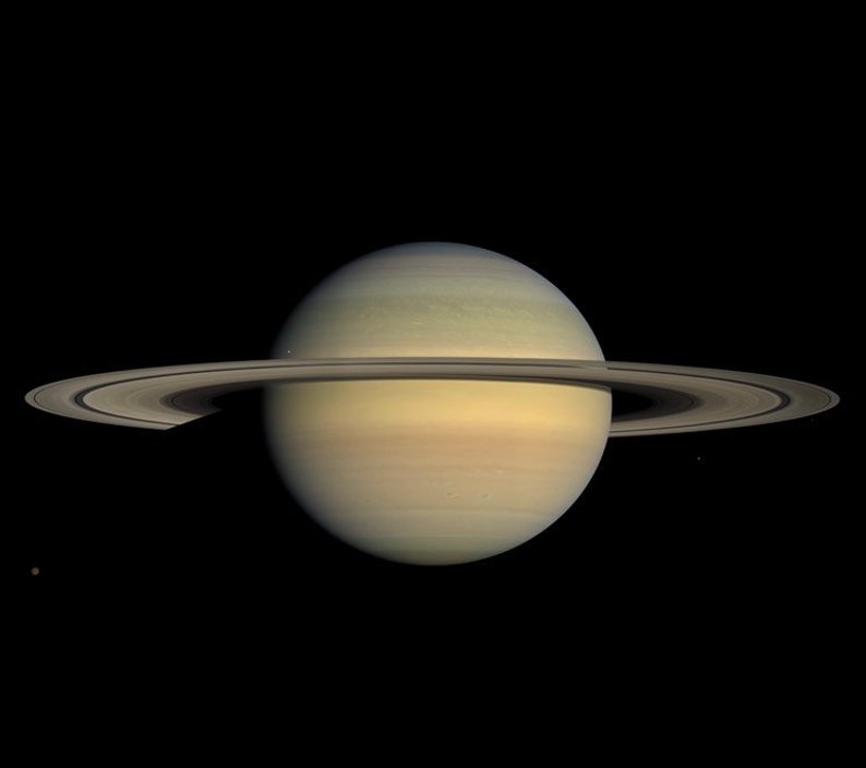 May include: An image of Saturn, a pale yellow planet with visible bands, set against a black backdrop. The planet is encircled by a prominent ring system. A small moon is visible to the lower left of the planet.