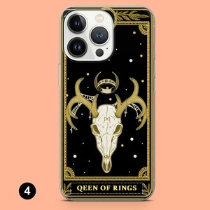 May include: A clear phone case with a black and gold design. The design features a skull with antlers, a crescent moon, and a ring. The text "QUEEN OF RINGS" is at the bottom of the design.