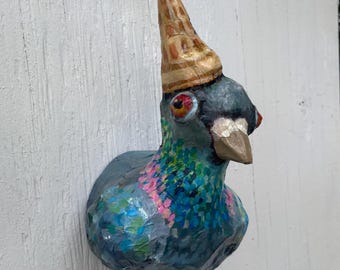 Può includere: Una scultura stravagante, dipinta a mano, di un piccione con un cappello a forma di cono. L'uccello è principalmente blu con accenti rosa e verdi iridescenti. Il cappello è color camoscio con strisce marroni. La scultura è montata su una superficie bianca.