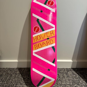 May include: A pink hoverboard with a striking design. It features diagonal white and green stripes, yellow accents, and the words "HOVER BOARD" in orange. Two red circles with the word "MATTEL" are also visible.