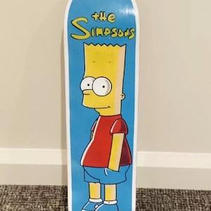 May include: A blue and white skateboard featuring a yellow cartoon character of Bart Simpson from The Simpsons. The text "The Simpsons" is printed in yellow on the board.