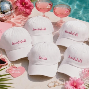 Puede incluir: Cinco gorras de béisbol blancas con texto bordado. Cuatro gorras presentan la palabra bombshell en hilo rosa, y una gorra presenta la palabra bride en hilo rosa. Las gorras están dispuestas con accesorios rosas, incluyendo gafas de corazón.
