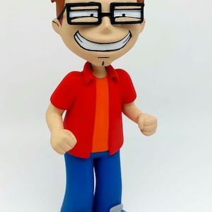 Op de afbeelding: Een handgeschilderd beeldje van een cartoonfiguur dat een rood shirt, een blauwe spijkerbroek en een bril draagt. Het personage heeft bruin haar en een brede glimlach.