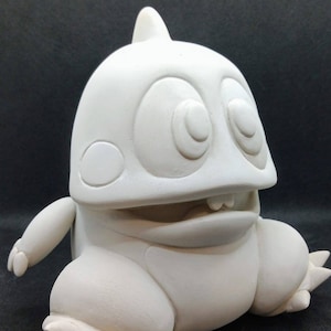 Könnte beinhalten: Weiße Keramikfigur eines Cartoon-Monsters mit großen Augen, einem breiten Mund und einem zahnigen Grinsen. Das Monster hat einen runden Körper und kurze, stummelige Arme und Beine.