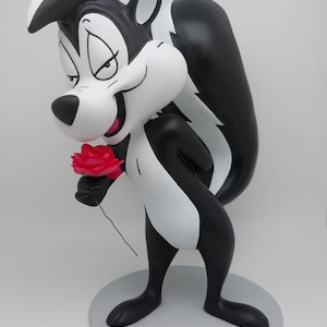Puede incluir: Una figurita de mofeta de dibujos animados en blanco y negro sosteniendo una rosa roja. La figurita está de pie sobre una base gris.