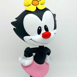 Puede incluir: Un personaje de dibujos animados en blanco y negro con una falda rosa y una flor amarilla en la cabeza. El personaje tiene una gran sonrisa y está de pie con las manos en las caderas.