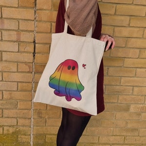 May include: A white tote bag with a rainbow ghost design. The ghost has a heart above its head. The bag is being held by a person wearing a brown sweater and a burgundy dress.
