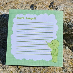 Op de afbeelding: Een vierkant, lichtgroen notitieblok met een witte, wolk-vormige rand en gelijnd papier. De tekst "Don't forget!" staat bovenaan. Een groen cartoonfiguur wijst naar het gelijnde gebied. Het notitieblok ligt op een rots.