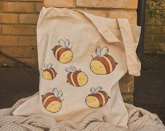 May include: A beige canvas tote bag with a brown handle and six cartoon bees printed on the front. The bees are yellow and brown with black stripes and have smiling faces.