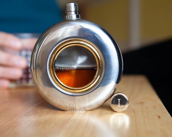 May include: A silver flask with a round, flat design and a gold-rimmed window revealing a brown liquid inside. The flask has a small, round, gold-colored cap with the letter "L" engraved on it.