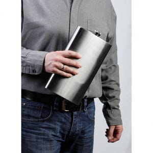 May include: A stainless steel hip flask with a hinged cap. The flask is rectangular and has a brushed metal finish. The person is wearing a grey button-down shirt and blue jeans. The flask is held in the person's hand.