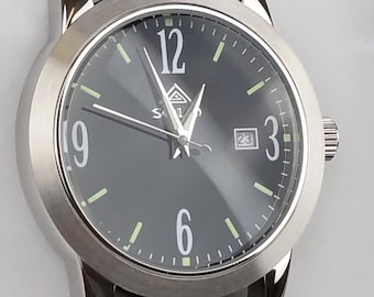 May include: A silver wristwatch with a black face and black leather band. The watch has luminous hands and hour markers, a date window at the 3 o'clock position, and a silver logo with the text "S.L." on the face.