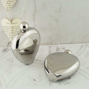 Op de afbeelding: Twee zilveren hartvormige hipflasks. De flasks zijn gemaakt van roestvrij staal en hebben een glanzende afwerking.
