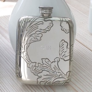 Puede incluir: Un frasco de cadera de plata con un diseño floral grabado y el nombre "GAH" en la parte delantera.