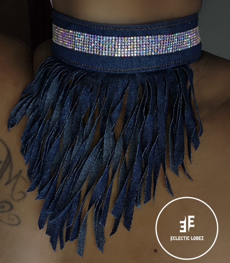 May include: A denim choker necklace with a fringe trim and a row of iridescent rhinestones. The choker is blue and the rhinestones are clear. The brand name "Eclectic Lobez" is printed in white on a black circle.