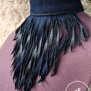 May include: A denim choker necklace with a fringe trim. The fringe is made of denim and is about 3 inches long. The choker is a dark blue color and has a brown stitching detail. The choker is displayed on a purple fabric background.