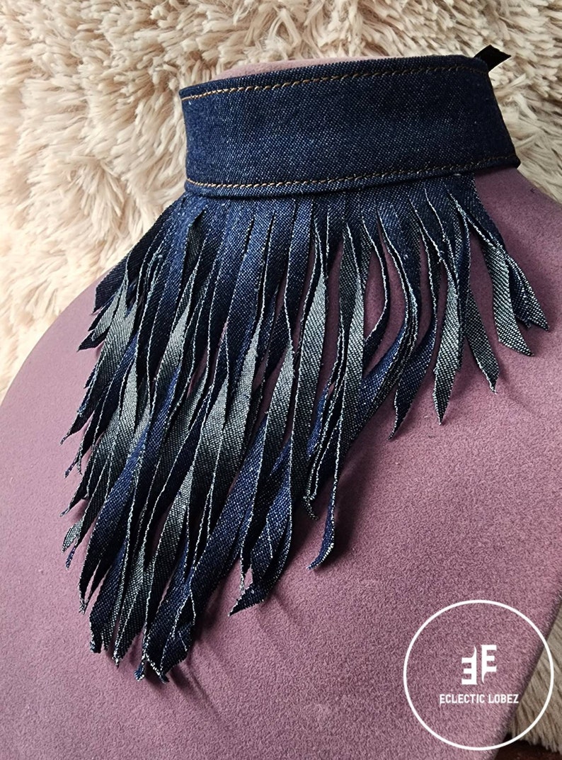 May include: A denim choker necklace with a fringe trim. The fringe is made of denim and is about 3 inches long. The choker is a dark blue color and has a brown stitching detail. The choker is displayed on a purple fabric background.