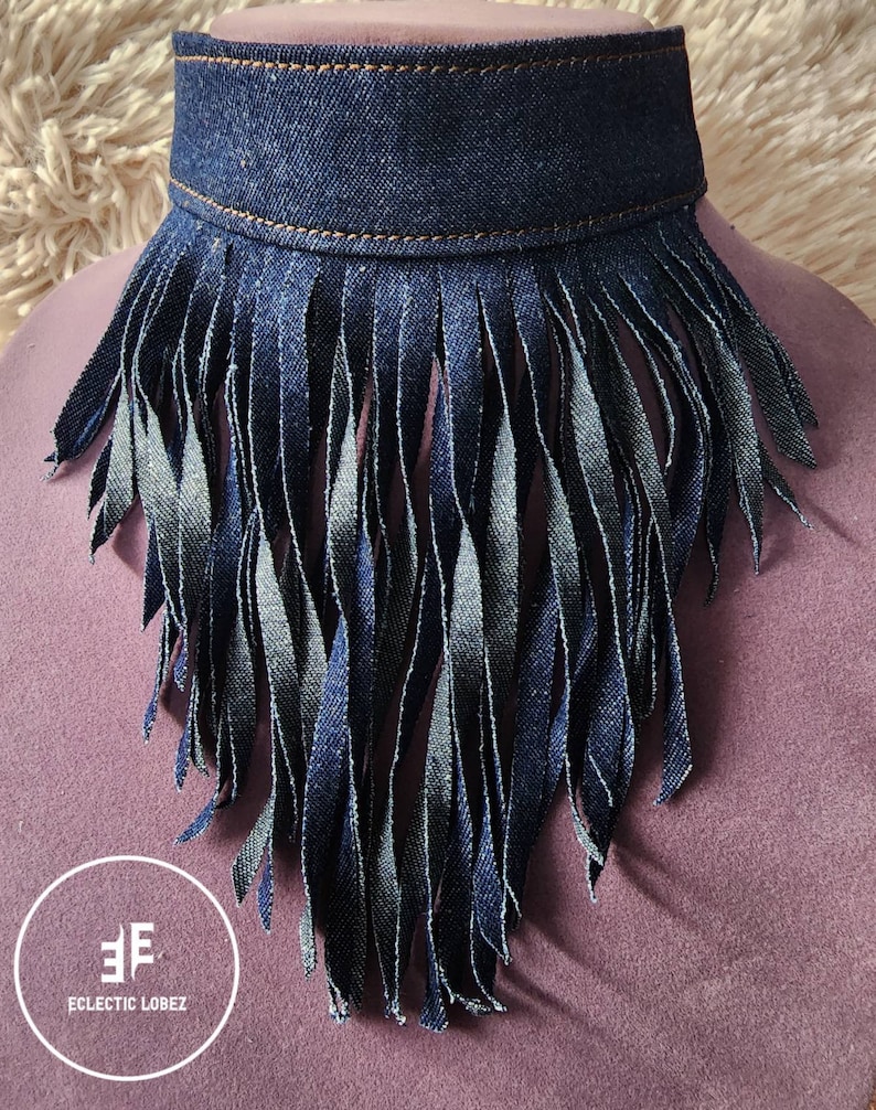 May include: A denim choker necklace with a fringe trim. The fringe is made of denim and is about 3 inches long. The choker is a dark blue color and has a brown stitching detail.