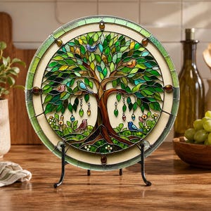 May include: A circular stained glass suncatcher featuring a tree of life design with green leaves, brown branches, and small colorful birds perched on the limbs. The piece is framed in a green border and displayed on a black metal stand.