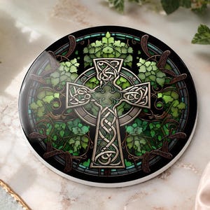 A round ceramic coaster with a Celtic cross design in shades of green and brown, surrounded by ivy-like foliage, on a marble surface with a white rose and green leaves.