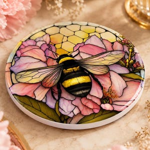 A circular ceramic coaster featuring a stained-glass style illustration of a bumblebee on pink flowers against a yellow honeycomb background.