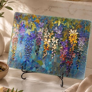 A rectangular glass cutting board with a stained-glass wisteria design stands on a black metal display stand on a white marble counter, with a cheese board nearby.