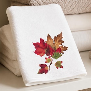 May include: A white hand towel featuring a printed illustration of autumn maple leaves in shades of red, orange, and green. The towel is folded and placed among a stack of neutral-colored towels, a beige ceramic vase, and a soap dispenser.