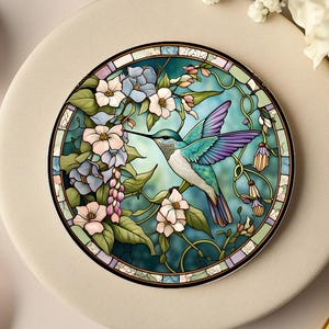 Puede incluir: Un posavasos redondo con un diseño de colibrí estilo vidriera. El colibrí tiene alas turquesas y moradas, un largo pico negro, y está rodeado de flores en tonos rosas, azules y blancos. El posavasos mide aproximadamente 10 cm de diámetro.