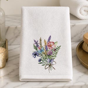 May include: White hand towel with a printed floral bouquet design. The bouquet features pink, purple, blue, and green flowers tied with a brown ribbon. The towel is displayed on a marble countertop.