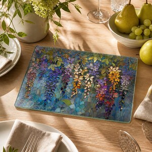 A rectangular glass cutting board with a stained-glass wisteria design is centered on a wooden table set with plates, glasses, and a bowl of fruit.