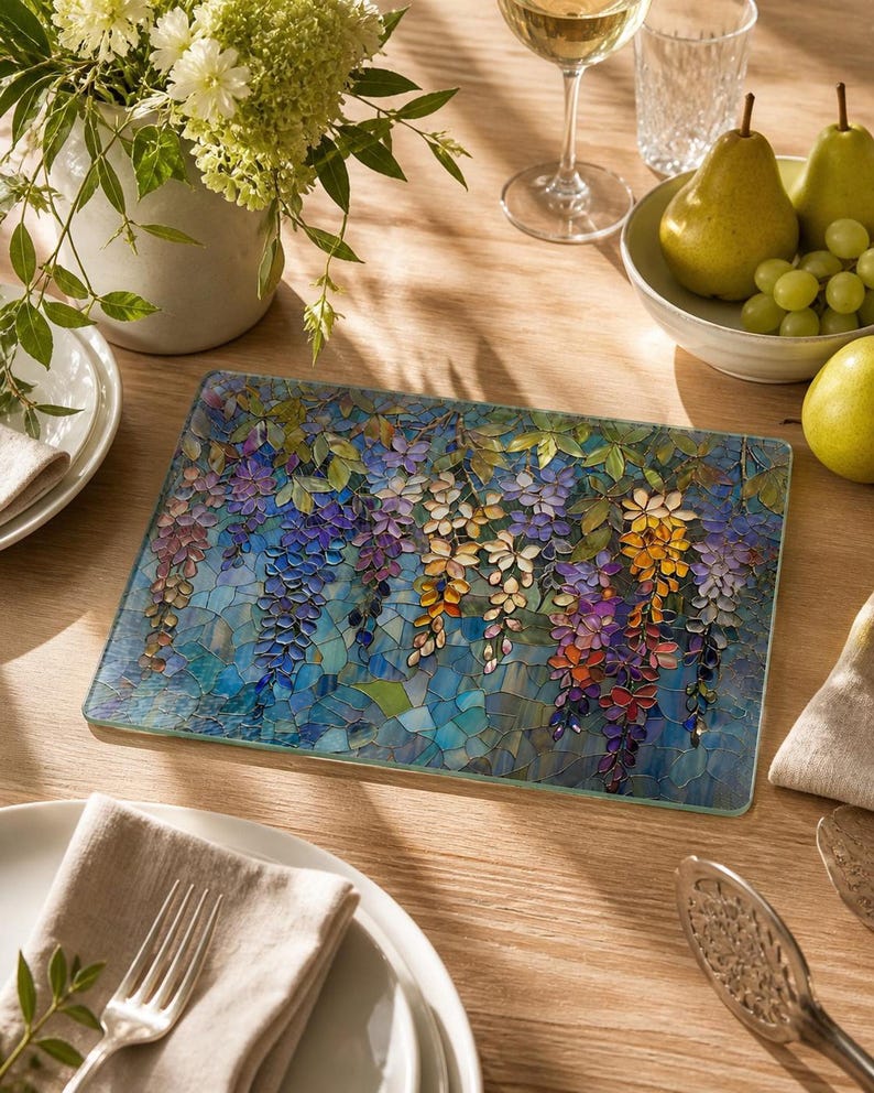 A rectangular glass cutting board with a stained-glass wisteria design is centered on a wooden table set with plates, glasses, and a bowl of fruit.
