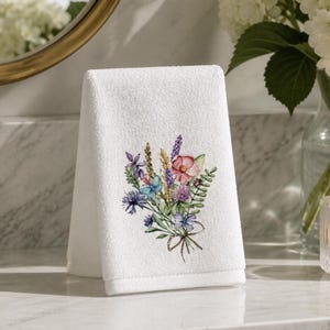 May include: A white hand towel featuring a printed watercolor illustration of a wildflower bouquet. The floral arrangement includes pink, purple, and blue blossoms, green foliage, and two small blue butterflies, tied together with a thin brown string.