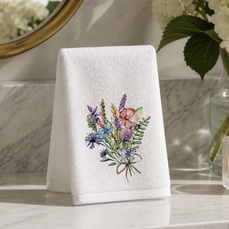 May include: A white hand towel featuring a printed watercolor illustration of a wildflower bouquet. The floral arrangement includes pink, purple, and blue blossoms, green foliage, and two small blue butterflies, tied together with a thin brown string.