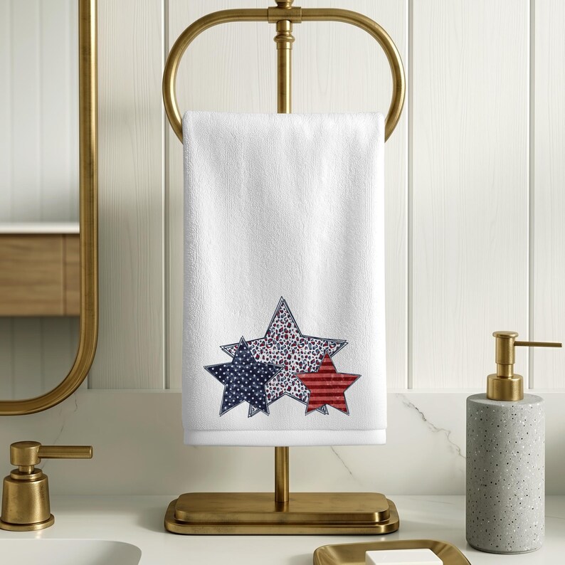 May include: White hand towel with three star designs in red, white, and blue. The stars are decorated with polka dots, stripes, and floral patterns. The towel is displayed on a gold-colored towel rack.