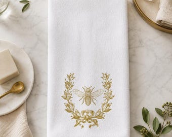 May include: A white rectangular hand towel featuring a centered gold-colored embroidered design of a bee surrounded by a laurel wreath and a ribbon bow at the base. The fabric has a soft, plush texture and clean, finished edges on all sides.