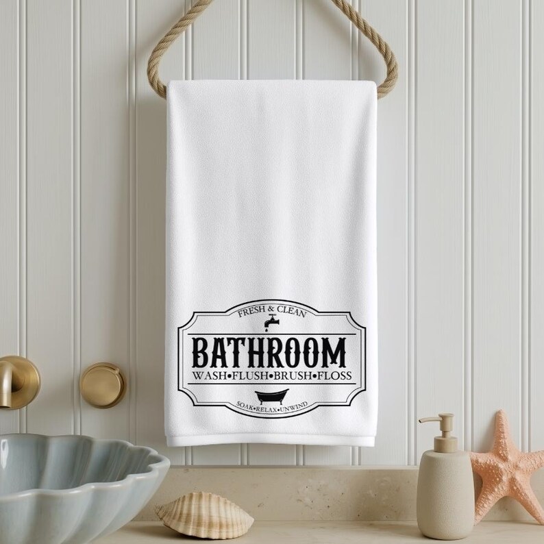May include: A white rectangular hand towel hanging from a rope, featuring a black graphic print with the text BATHROOM, WASH, FLUSH, BRUSH, FLOSS, FRESH & CLEAN, and SOAK, RELAX, UNWIND, along with icons of a water faucet and a bathtub.