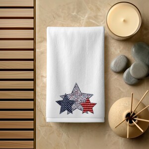 May include: White hand towel with three star designs in red, white, and blue. The stars have different patterns, including polka dots and stripes. The towel is on a light brown surface with a candle and decorative items.