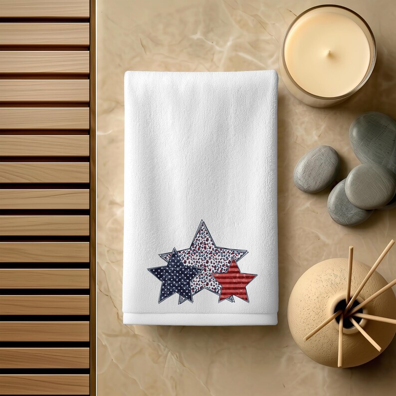 May include: White hand towel with three star designs in red, white, and blue. The stars have different patterns, including polka dots and stripes. The towel is on a light brown surface with a candle and decorative items.