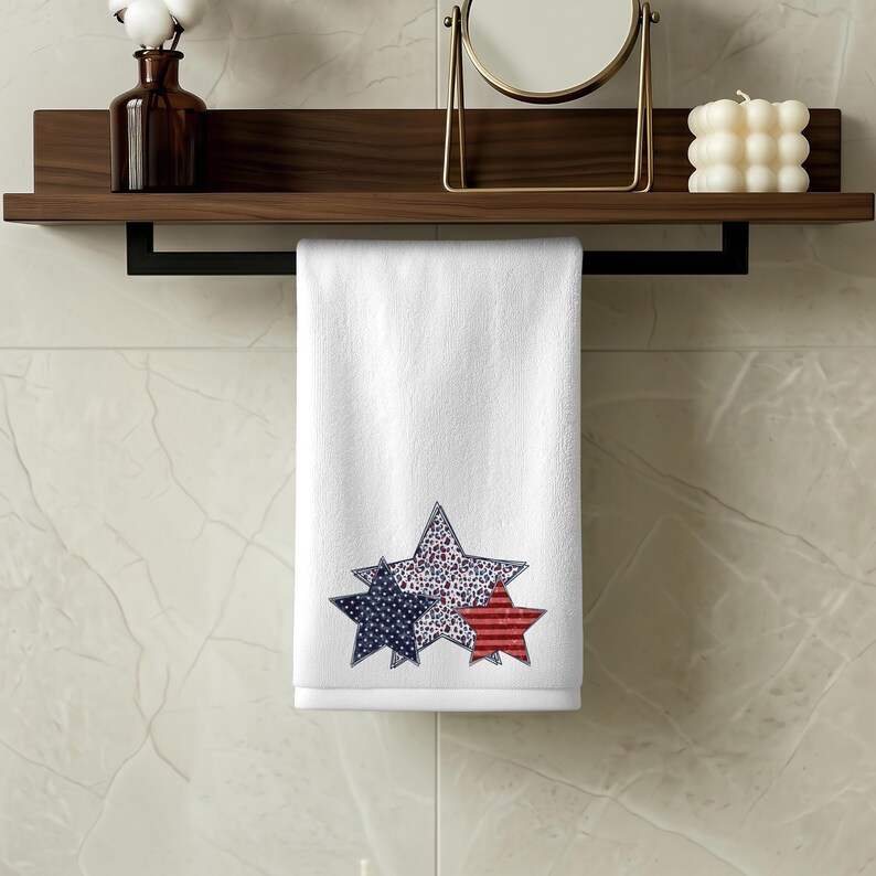 May include: White hand towel with three star designs in red, white, and blue. The stars have different patterns: one with red stripes, one with blue dots, and one with a mix of red and white dots. The towel is hanging on a black bar.