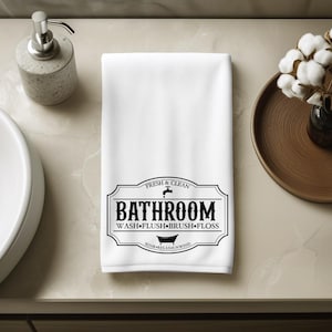 May include: A white rectangular hand towel featuring a black graphic print in the center. The design includes a decorative border, a faucet icon, a bathtub icon, and the text: FRESH & CLEAN, BATHROOM, WASH FLUSH BRUSH FLOSS, SOAK RELAX UNWIND.