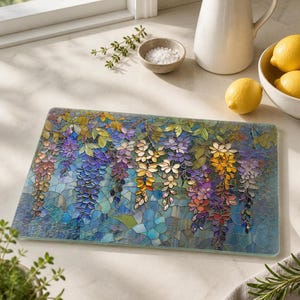 A rectangular glass cutting board with a stained-glass wisteria design rests on a white counter next to a white pitcher, a bowl of lemons, and salt.
