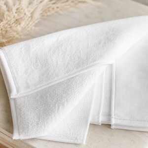 May include: Folded white towel with a textured surface and a stitched border. The towel is made of a soft material and is folded to display multiple layers. The image is well-lit, highlighting the clean, crisp appearance of the towel.