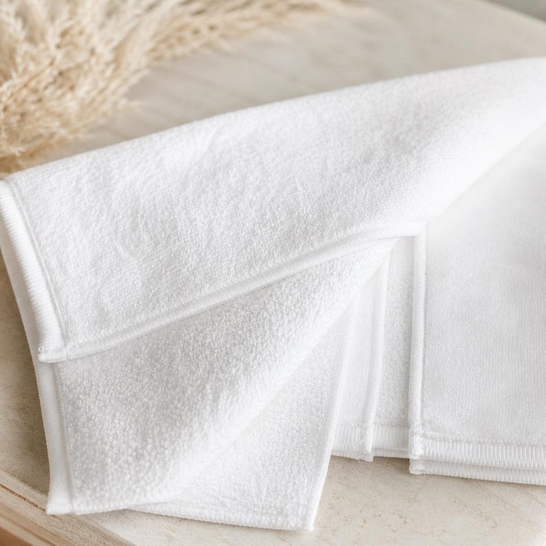 May include: Folded white towel with a textured surface and a stitched border. The towel is made of a soft material and is folded to display multiple layers. The image is well-lit, highlighting the clean, crisp appearance of the towel.