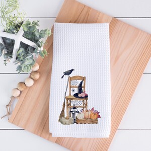 May include: A white waffle-weave kitchen towel featuring a printed illustration of a wooden chair, a black crow, a witch hat, a broom, a spider web, a lantern, and a collection of pumpkins and autumn leaves arranged in a wooden crate.