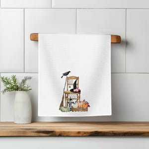 May include: A white waffle-textured kitchen towel featuring a watercolor illustration of a wooden chair, a black crow, a broom, a witch hat, a lantern, and a collection of pumpkins and autumn leaves arranged on the floor.