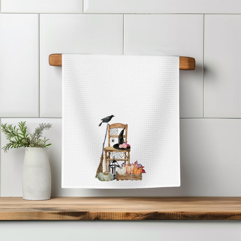 May include: A white waffle-textured kitchen towel featuring a watercolor illustration of a wooden chair, a black crow, a broom, a witch hat, a lantern, and a collection of pumpkins and autumn leaves arranged on the floor.
