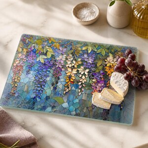 A rectangular glass cutting board with a stained-glass wisteria design holds slices of cheese and red grapes on a white marble counter.