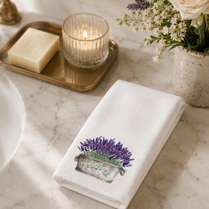 A white folded hand towel with a printed image of purple lavender flowers in a gray planter with "FLOWER" on it, on a marble countertop with a lit candle and a vase of white flowers.