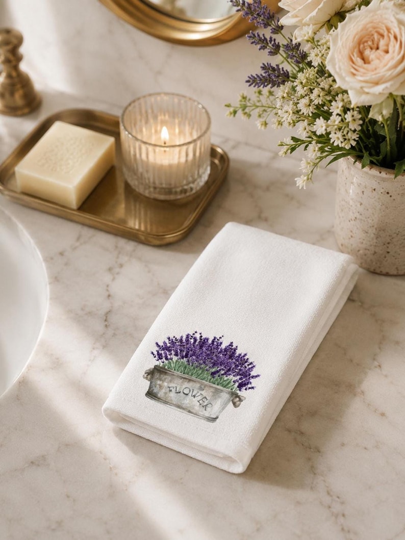 A white folded hand towel with a printed image of purple lavender flowers in a gray planter with "FLOWER" on it, on a marble countertop with a lit candle and a vase of white flowers.